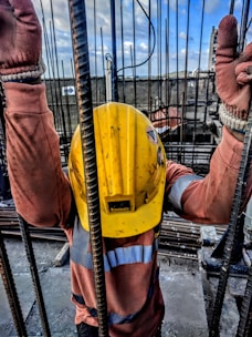 A construction worker wearing a yellow safety helmet and orange protective gloves is holding onto vertical metal rods. The person is dressed in a long-sleeved high-visibility shirt. The scene appears to be on a construction site with a cloudy sky and structural elements visible in the background.