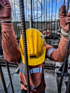 A construction worker wearing a yellow safety helmet and orange protective gloves is holding onto vertical metal rods. The person is dressed in a long-sleeved high-visibility shirt. The scene appears to be on a construction site with a cloudy sky and structural elements visible in the background.