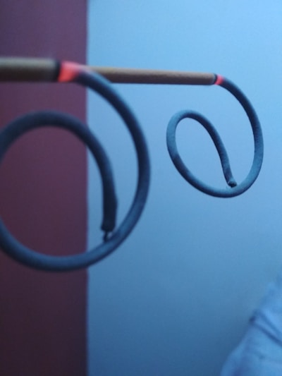 Two spiral mosquito coils are suspended horizontally on a stick. They are partially burnt and the tips glow red. The background consists of a plain wall, partially visible in red and light blue.