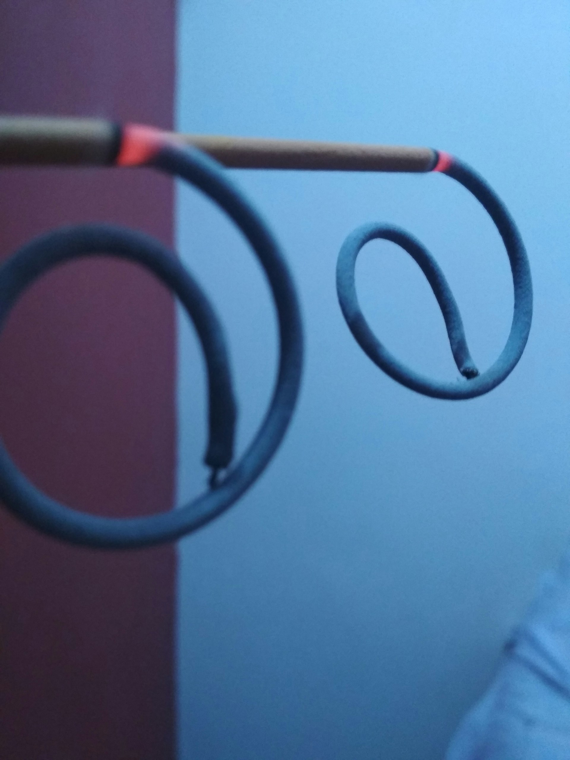 Two spiral mosquito coils are suspended horizontally on a stick. They are partially burnt and the tips glow red. The background consists of a plain wall, partially visible in red and light blue.