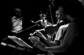 A black and white image captures a group of musicians playing instruments on stage. The focus is on three individuals playing guitar and violin, with expressive gestures suggesting engagement with their music. A man in the background stands near a music stand, adding depth to the scene.
