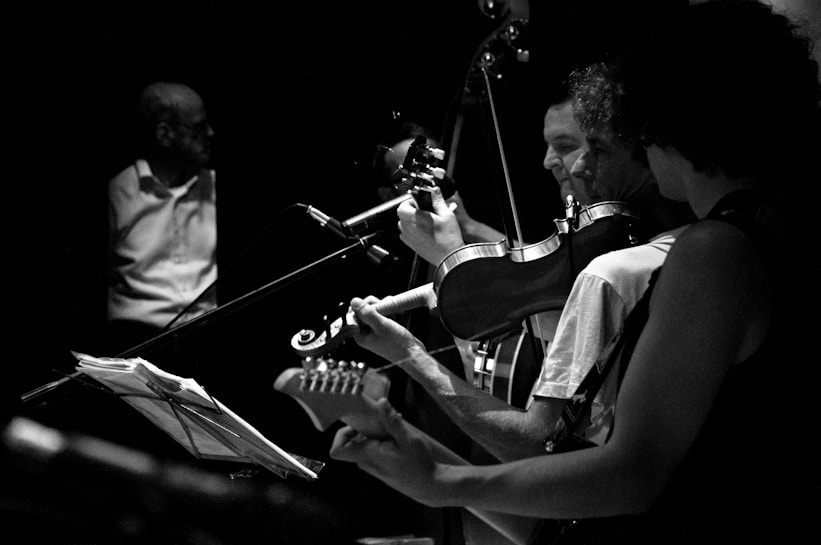 A black and white image captures a group of musicians playing instruments on stage. The focus is on three individuals playing guitar and violin, with expressive gestures suggesting engagement with their music. A man in the background stands near a music stand, adding depth to the scene.