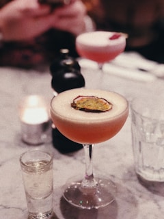 A sophisticated cocktail garnished with a slice of passion fruit is presented in an elegant stem glass on a marble table. Nearby, a shot glass filled with a clear liquid and a blurred background highlight other drinks and ambient lighting. The setting suggests a relaxed, upscale social environment.