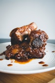 Succulent ribs coated in a rich, glossy barbecue sauce, resting on a white plate. The meat appears tender and well-cooked, with some sauce drizzled around the plate.
