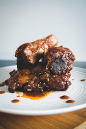 Succulent ribs coated in a rich, glossy barbecue sauce, resting on a white plate. The meat appears tender and well-cooked, with some sauce drizzled around the plate.