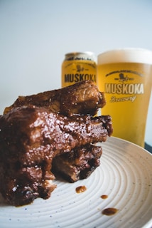 A plate holding a stack of glazed barbecue ribs is in the foreground. Behind the ribs, there is a glass and a can of beer from Muskoka Brewery, both filled. The plate is white and speckled, and the beer has a foamy head.