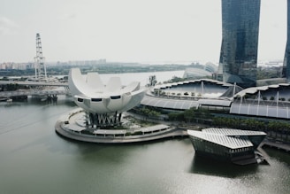 A modern cityscape featuring an innovative building with a lotus flower shape surrounded by water. The structure is adjacent to a large, curved architectural complex near a tall skyscraper. In the background, there's a large ferris wheel and distant urban landscape. Lush greenery is visible around the buildings, which gives a sense of harmony between nature and urban design.