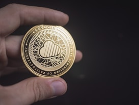 A hand is holding a shiny, gold-colored coin with intricate designs and the word 'Skycoin' inscribed on it. The coin features a cloud emblem in the center with a network-like pattern surrounding it, suggesting a technological or digital theme.