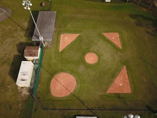 Aerial view of a baseball or softball infield with clearly visible bases and pitcher's mound. The surrounding area includes a building, storage container, and light poles. The infield is made of reddish clay, while the outfield is grass. Shadows from the light poles and structures are cast onto the field.