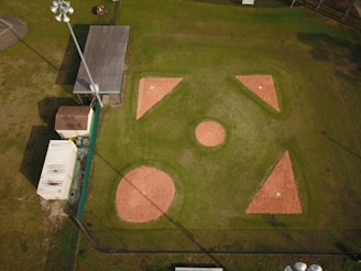 Aerial view of a baseball or softball infield with clearly visible bases and pitcher's mound. The surrounding area includes a building, storage container, and light poles. The infield is made of reddish clay, while the outfield is grass. Shadows from the light poles and structures are cast onto the field.
