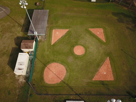 Aerial view of a baseball or softball infield with clearly visible bases and pitcher's mound. The surrounding area includes a building, storage container, and light poles. The infield is made of reddish clay, while the outfield is grass. Shadows from the light poles and structures are cast onto the field.