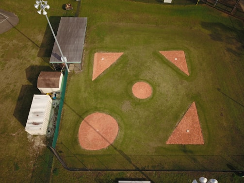 Aerial view of a baseball or softball infield with clearly visible bases and pitcher's mound. The surrounding area includes a building, storage container, and light poles. The infield is made of reddish clay, while the outfield is grass. Shadows from the light poles and structures are cast onto the field.