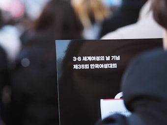 A close-up view of a dark placard featuring Korean text, surrounded by blurred figures in the background, suggesting a public event or gathering.