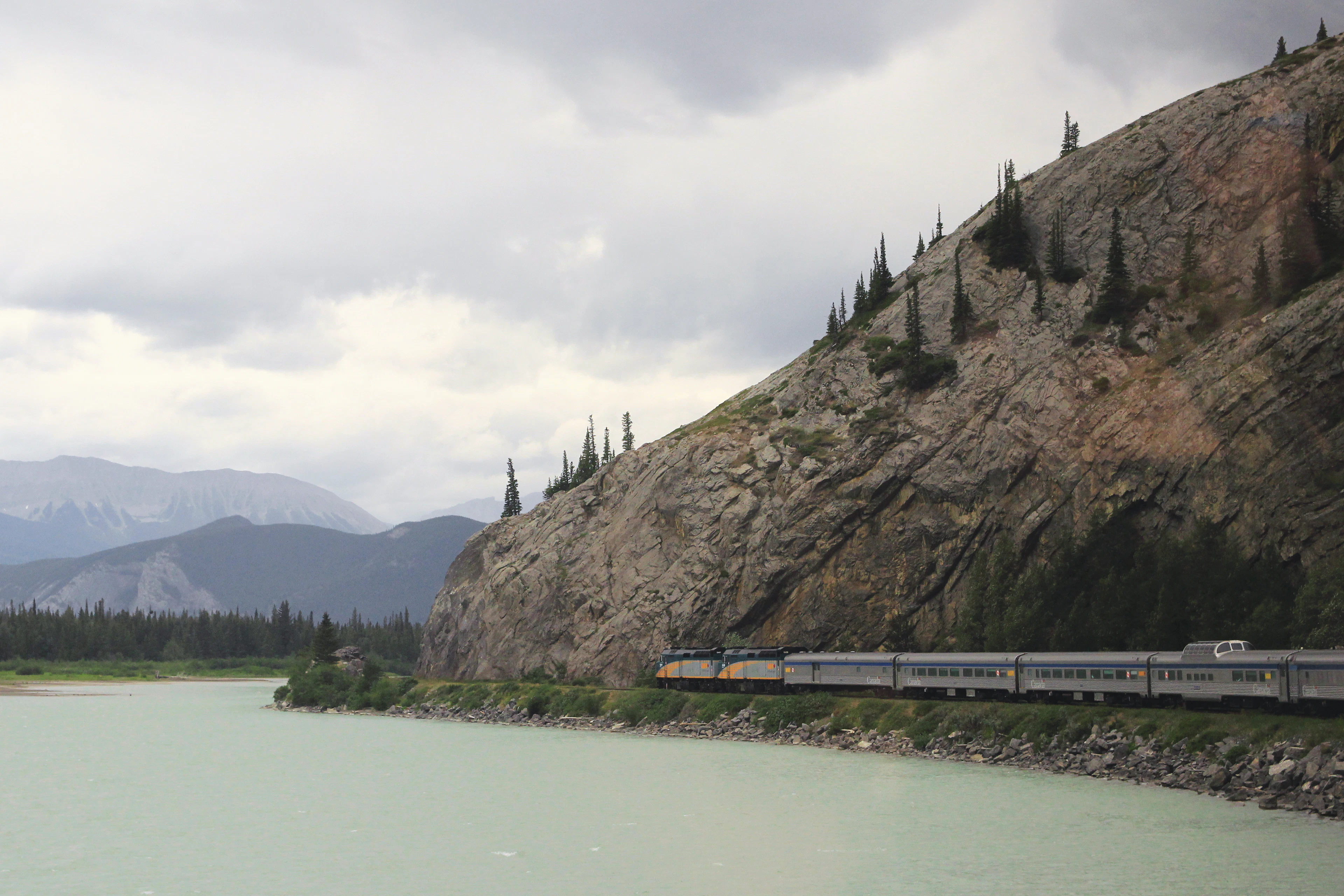 A passenger train curving beneath a rocky slope beside the water in the Rockies near Jasper