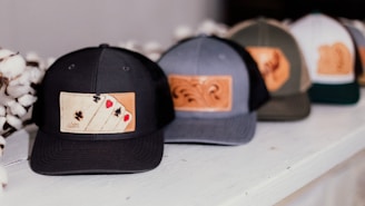 A row of baseball caps featuring leather patches with different designs, including one with playing card suits. The caps are placed on a white surface, and cotton flowers are visible beside them.