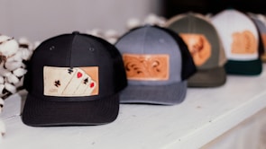 A row of baseball caps featuring leather patches with different designs, including one with playing card suits. The caps are placed on a white surface, and cotton flowers are visible beside them.