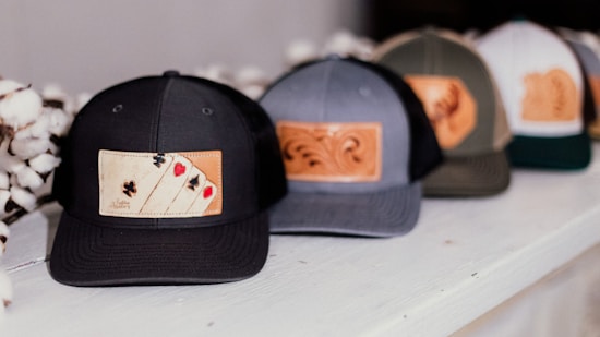 A row of baseball caps featuring leather patches with different designs, including one with playing card suits. The caps are placed on a white surface, and cotton flowers are visible beside them.
