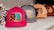 A row of colorful caps placed on a white surface. The closest cap is bright pink with a patch featuring a blue rose and leopard print design. The other caps are in shades of gray, peach, and maroon, each with different decorative patches.