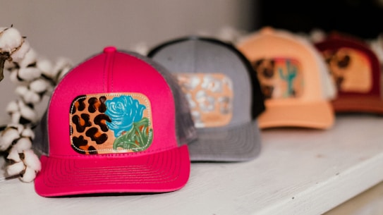 A row of colorful caps placed on a white surface. The closest cap is bright pink with a patch featuring a blue rose and leopard print design. The other caps are in shades of gray, peach, and maroon, each with different decorative patches.