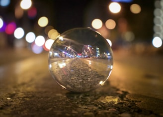 A crystal ball on a gravel path reflects the surrounding urban nightscape, with colorful bokeh lights out of focus in the background creating a dreamy atmosphere.