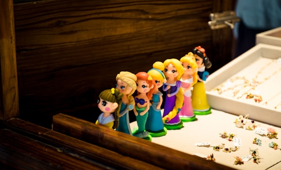 A row of colorful, cartoonish princess figurines lined up on a beige surface, with wooden surroundings. These figurines stand near a display of intricate jewelry pieces.