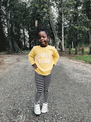 A young child stands confidently on a gravel path, surrounded by tall trees. The child is wearing a bright yellow sweatshirt with the words 'Girls Club' printed in white, black and white striped leggings, and white shoes. The background features a lush forest setting with dense greenery.