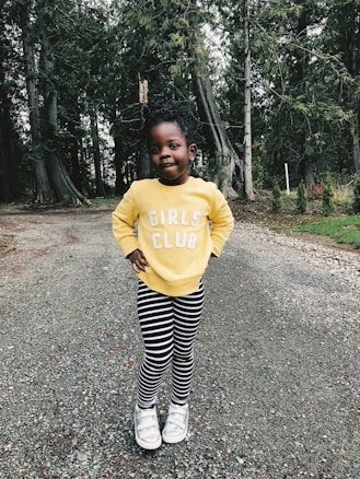 A young child stands confidently on a gravel path, surrounded by tall trees. The child is wearing a bright yellow sweatshirt with the words 