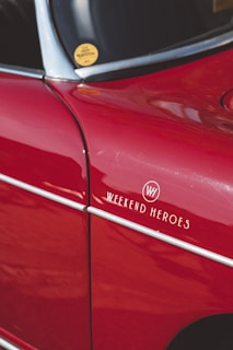 A close-up view of a vintage car that is predominantly red with chrome accents. There is a sticker or emblem on the car reading 'Weekend Heroes' and a small round yellow sticker on the window. The surface of the car reflects light, showing the smoothness and shine of the paint.
