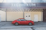 A vintage red sports car is parked in front of a building with a sign reading 'TECHNIKUM'. The structure has a retro design with a row of round light bulbs beneath the sign. There are three doors on the front of the building, with a neutral tone wall.