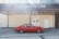 A vintage red sports car is parked in front of a building with a sign reading 'TECHNIKUM'. The structure has a retro design with a row of round light bulbs beneath the sign. There are three doors on the front of the building, with a neutral tone wall.