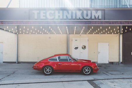 A vintage red sports car is parked in front of a building with a sign reading 'TECHNIKUM'. The structure has a retro design with a row of round light bulbs beneath the sign. There are three doors on the front of the building, with a neutral tone wall.