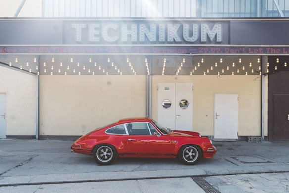 A vintage red sports car is parked in front of a building with a sign reading 'TECHNIKUM'. The structure has a retro design with a row of round light bulbs beneath the sign. There are three doors on the front of the building, with a neutral tone wall.