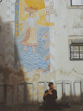 A mural on a weathered wall depicts a person carrying a basket on their head, surrounded by decorative patterns in blue, yellow, and orange tones. Below the mural, a man sits cross-legged playing a wooden flute.