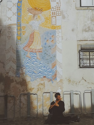 A mural on a weathered wall depicts a person carrying a basket on their head, surrounded by decorative patterns in blue, yellow, and orange tones. Below the mural, a man sits cross-legged playing a wooden flute.