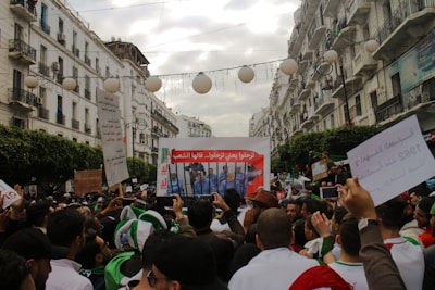 A large group of people is gathered in a street lined with white buildings, holding signs and banners in Arabic, with trees and lamp decorations overhead. The scene suggests a protest or a demonstration, with many individuals wearing caps and some holding flags.