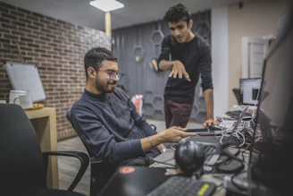 standing man next to sitting man in front of computer in room