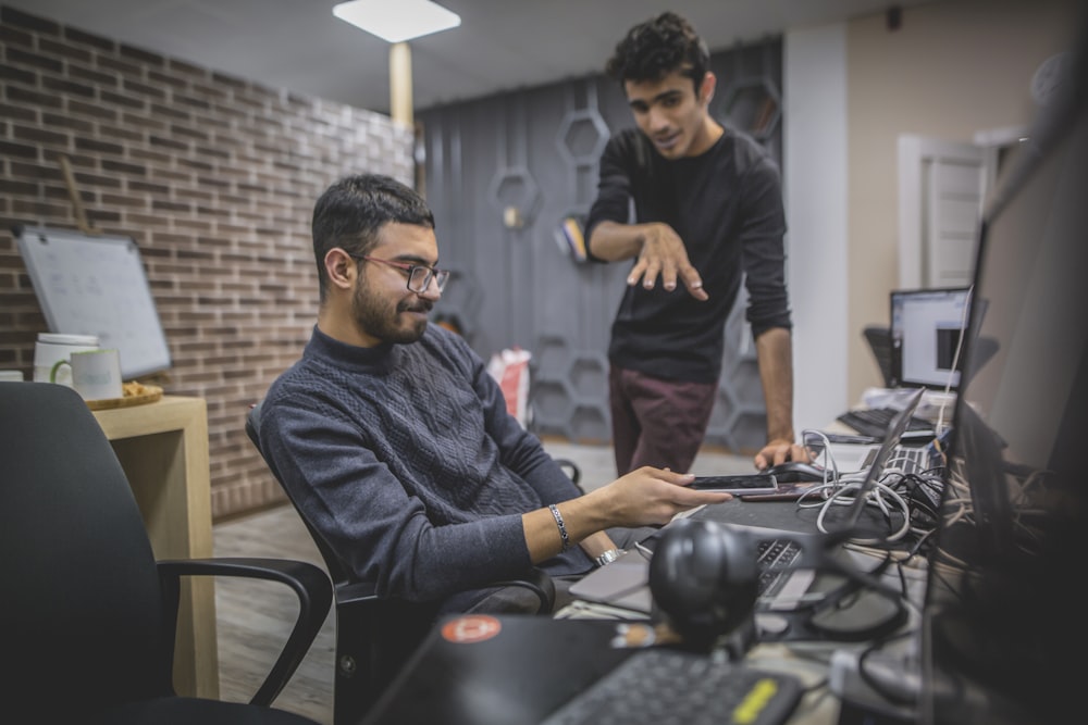 standing man next to sitting man in front of computer in room