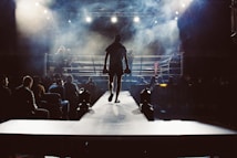 A boxer walks toward a well-lit ring through a path lined with spectators. The scene is filled with smoke and dramatic lighting, creating a theatrical atmosphere.