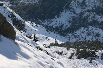 A snowy mountain landscape with several people visible mid-action, appearing excited or engaged, on a steep snow-covered slope. The background shows a rugged mountain view with patches of snow and trees.