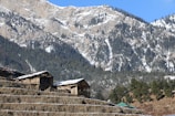 Wooden structures, possibly cabins or houses, sit atop terraced land with a backdrop of snow-dusted, forested mountains under a clear blue sky.