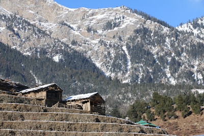 Wooden structures, possibly cabins or houses, sit atop terraced land with a backdrop of snow-dusted, forested mountains under a clear blue sky.