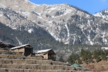 Wooden structures, possibly cabins or houses, sit atop terraced land with a backdrop of snow-dusted, forested mountains under a clear blue sky.