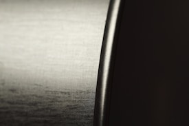 Close-up view of a shiny metallic surface with a smooth, curved edge. The texture has a brushed finish, reflecting light subtly across the surface. The lighting emphasizes the contrast between the polished metal and the darker background.