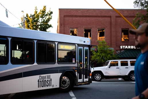 A public transport bus with 'Chapel Hill Transit' and the display message 'GO HEELS!' is passing by a street with a brick building housing a Starbucks. Parked beside the building is a white Jeep. A person in a baseball cap and blue shirt can be partially seen on the right side of the image.