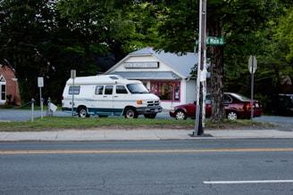 A small, white storefront with the sign 'Back Alley Bikes' surrounded by trees and parked vehicles, including a white van and a red car. A street sign indicates the intersection of Main Street, and a speed limit sign is visible. The scene is set in a cozy, suburban environment.
