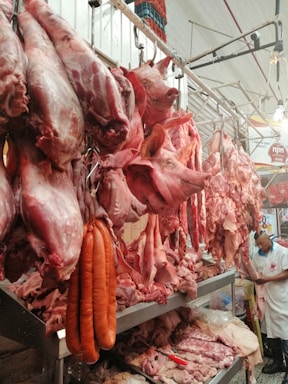 A butcher's shop displaying a variety of raw meats, including whole pig heads, hanging carcasses, and sausages. There is a man in a white uniform working behind the counter, surrounded by various cuts of meat on metal shelves.