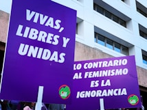 Two large purple signs with white text are held up by people. The text in Spanish promotes feminist messages, with one sign saying 'Vivas, Libres y Unidas' and the other stating 'Lo Contrario al Feminismo es la Ignorancia'. A building with large windows is visible in the background.