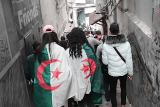 A group of people are walking down an urban alleyway that is narrow and lined with graffiti. Two individuals are draped in Algerian flags, displaying the national colors of green and white with a red star and crescent. The scene includes a variety of people wearing casual clothing, some of whom have their backs turned to the camera.