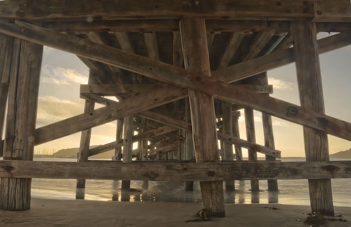 A perspective view under a wooden pier with structural beams crisscrossing in the foreground. The ocean and distant land can be seen through the gaps in the wood, with soft lighting suggesting either sunrise or sunset. The sandy beach beneath the pier reflects the wooden structure.