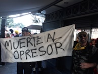 A group of people, some in costumes, are holding a large banner with the text 'MUERTE AL OPRESOR'. The scene appears to be set in an outdoor area with trees visible in the background and a covered walkway. One individual is wearing a cap labeled 'SWAT' and another person wearing sunglasses and a bandana is standing nearby. The mood is festive and possibly part of a protest or celebration.